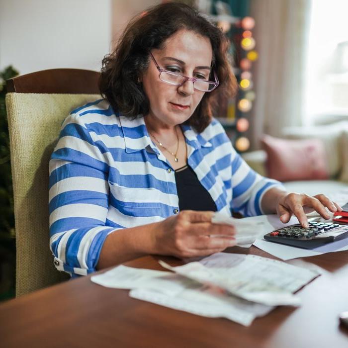 A woman in glasses adds up her receipts at a kitchen table.